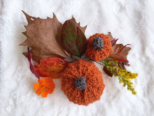 Two orange crocheted pumpkins with gray stems are arranged on a white textured blanket with autumn leaves, an orange flower, and yellow wildflowers.